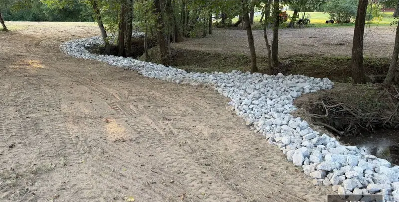 Creek bank lined with new riprap rock curving past a freshly graded slope under trees