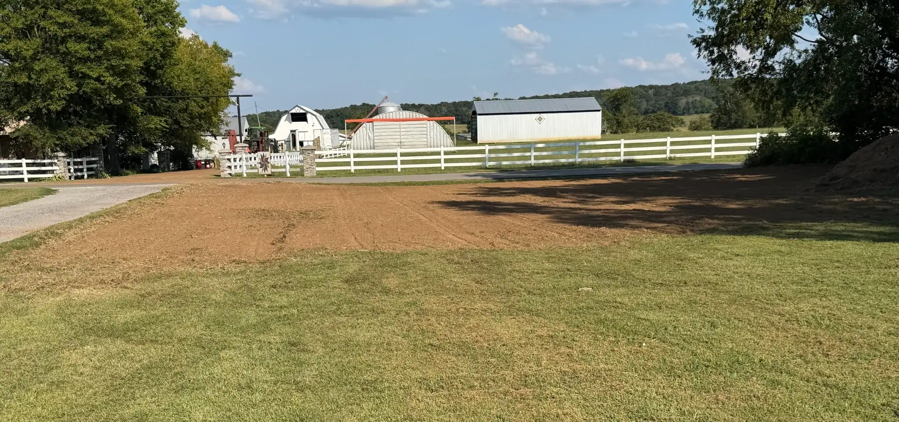 Wide view of a freshly graded yard beside white board fencing and barns