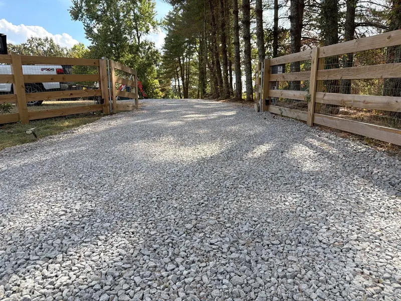 Freshly graded gravel driveway running through an open wooden farm gate under pine trees
