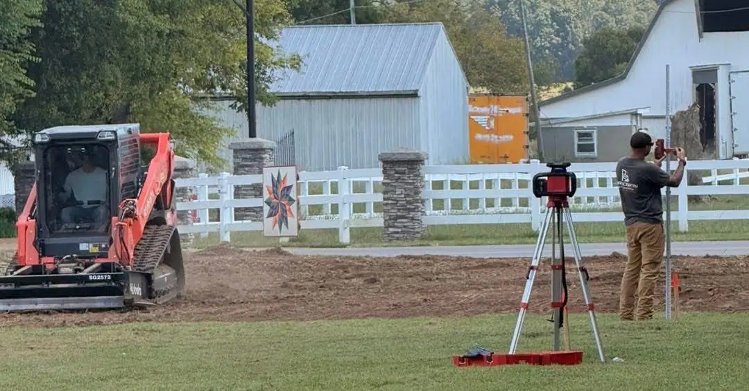 Operator reading a grade rod next to a laser level while a tracked loader works behind