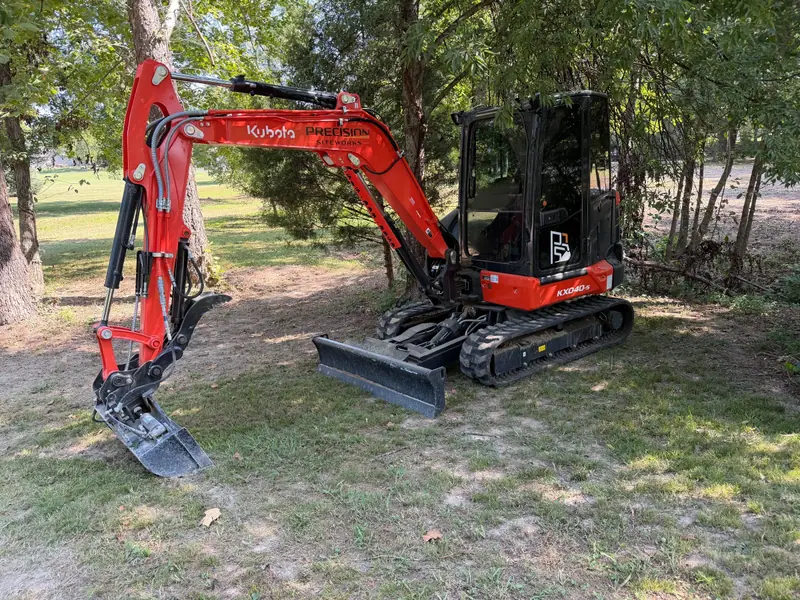 Orange Kubota compact excavator with a hydraulic thumb parked under shade trees