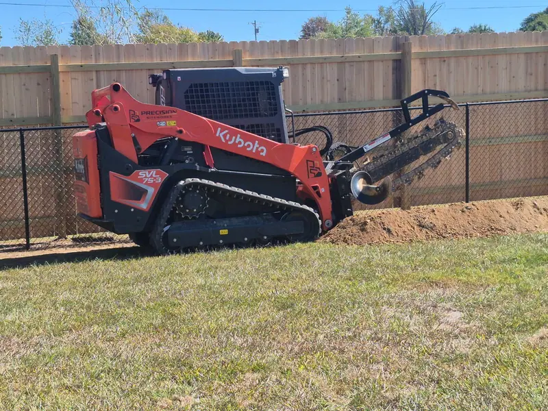 Orange Kubota tracked loader with a trencher attachment cutting a trench along a backyard fence